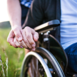 Close-up of a person in a wheelchair holding hands with someone standing beside them in an outdoor setting.