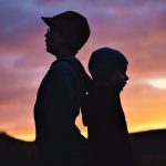 Silhouettes of two children standing back-to-back against a colorful sunset sky.
