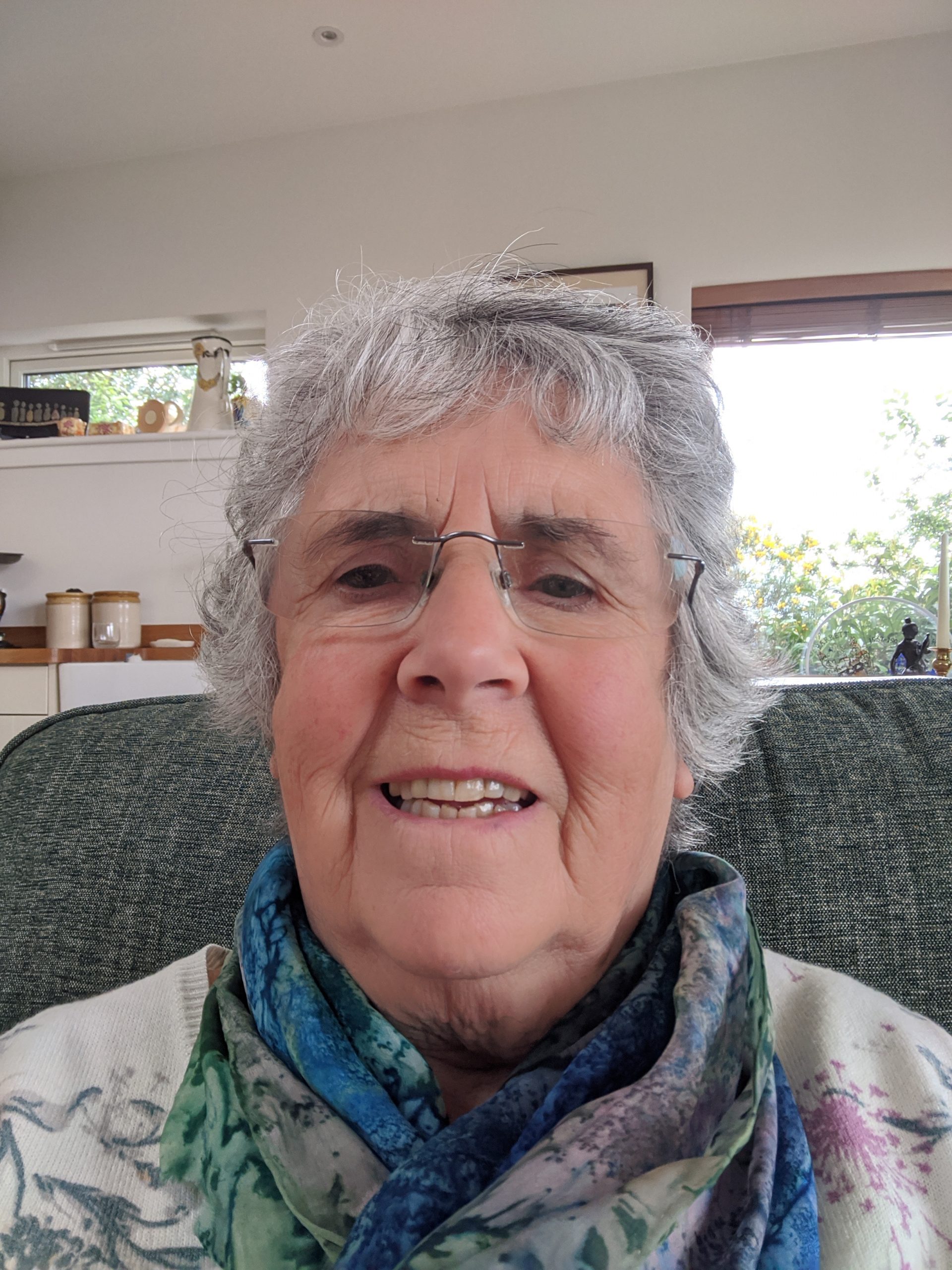 Older white lady with short grey hair and glasses sitting on a sofa wearing a flowery top.