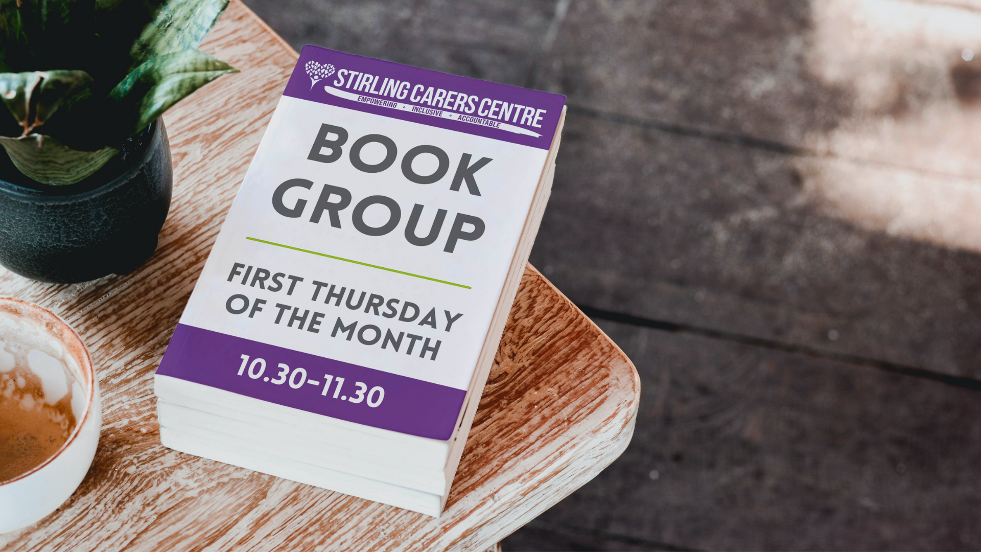 A stack of flyers on a wooden table promoting a Book Group hosted by Stirling Carers Centre. The text reads, Book Group - First Thursday of the Month - 10:30-11:30. A plant and a coffee cup with latte art are beside the flyers.