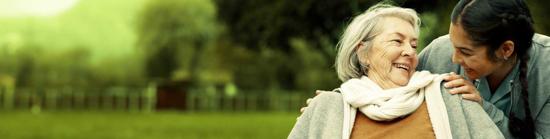 Elderly woman in wheelchair smiling and enjoying a moment with a caregiver in an outdoor park setting.