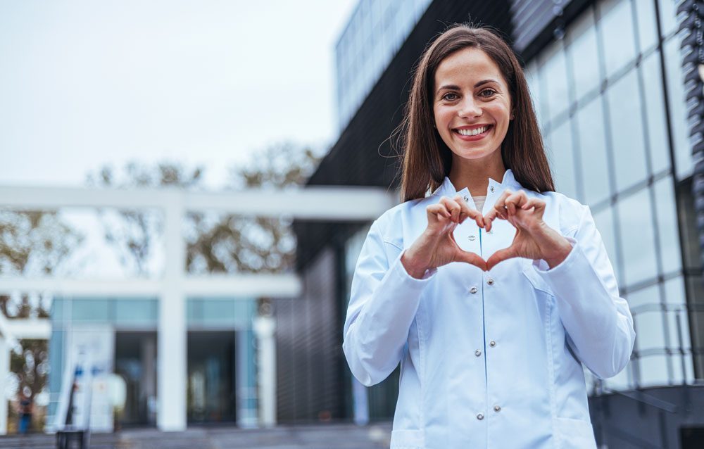 A smiling woman in a white lab coat stands in front of a modern building, forming a heart shape with her hands.