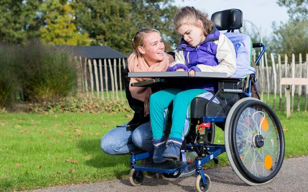 A woman crouches next to a young girl in a wheelchair outdoors. They are both smiling, and the woman appears to be engaging with the girl in conversation. The scene is set in a park with green grass, trees, and a wooden fence in the background.