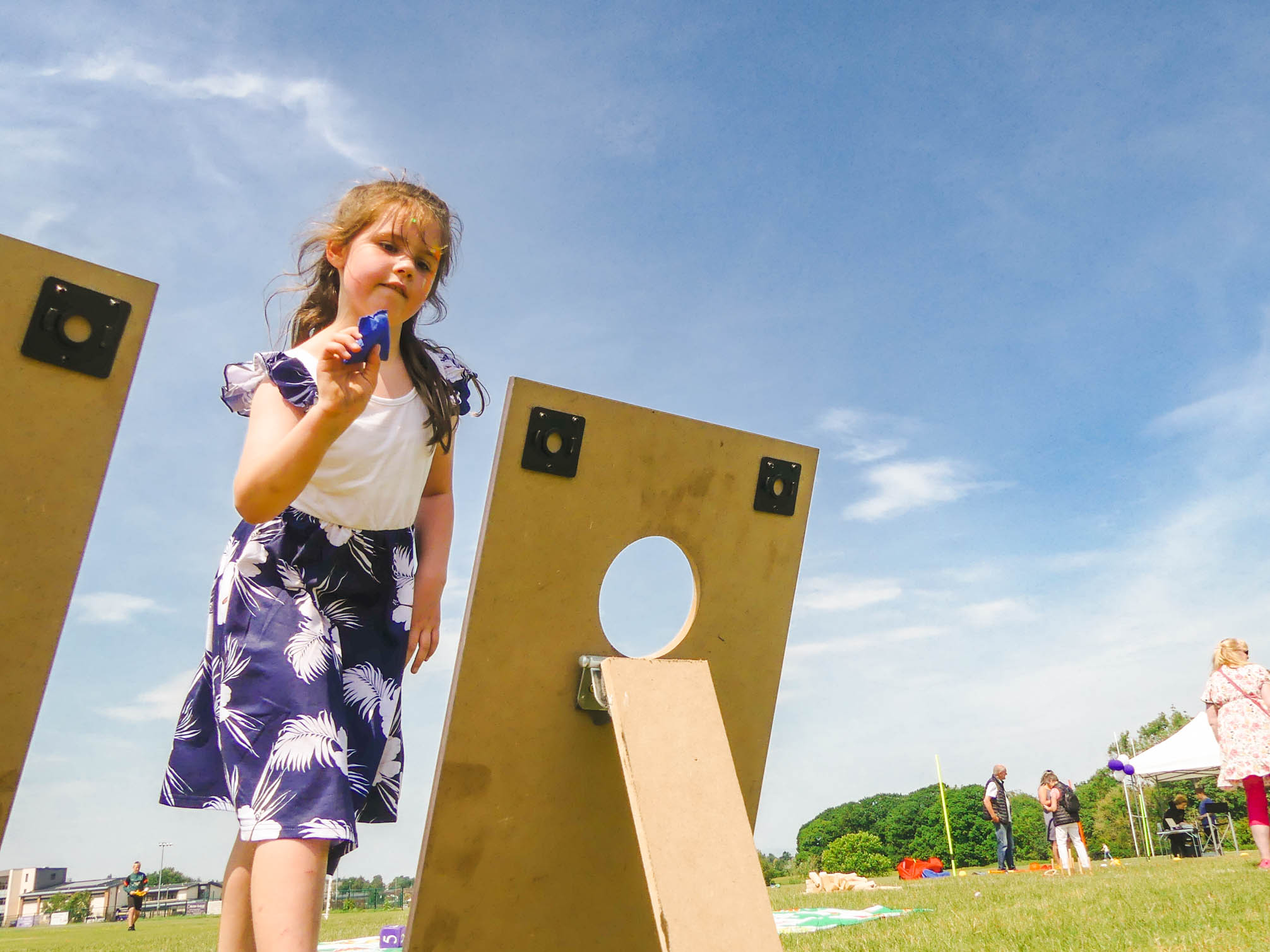 A young girl with light brown hair wearing a navy and white dress throws a blue bean bag through a wooden board with a circular hole during an outdoor event. The sky is clear and blue, and people are mingling in the background.