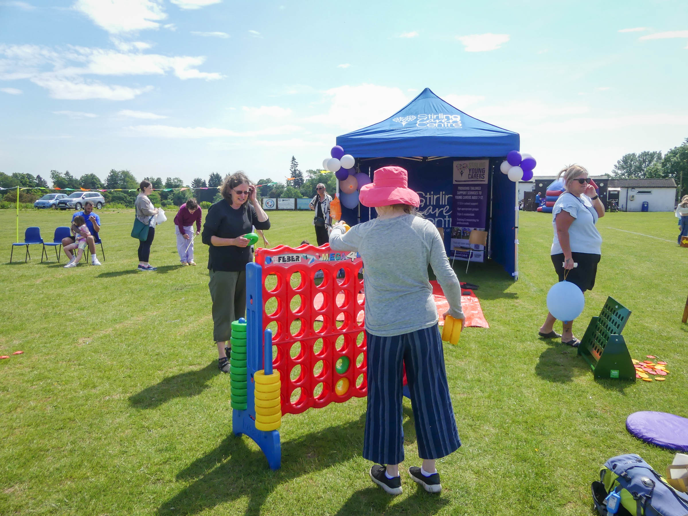 People gather outdoors on a grassy area under a sunny sky, participating in various activities. In the foreground, a person wearing a pink hat plays a large Connect Four game. A blue tent and other participants in the background suggest a community event.