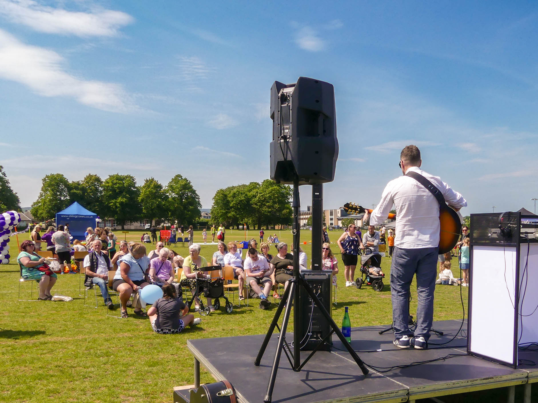 A musician stands on a stage, playing guitar and singing into a microphone at an outdoor event. A crowd of people, including families with children, sit on the grass, enjoying the performance. The sky is clear and blue, with trees in the background.