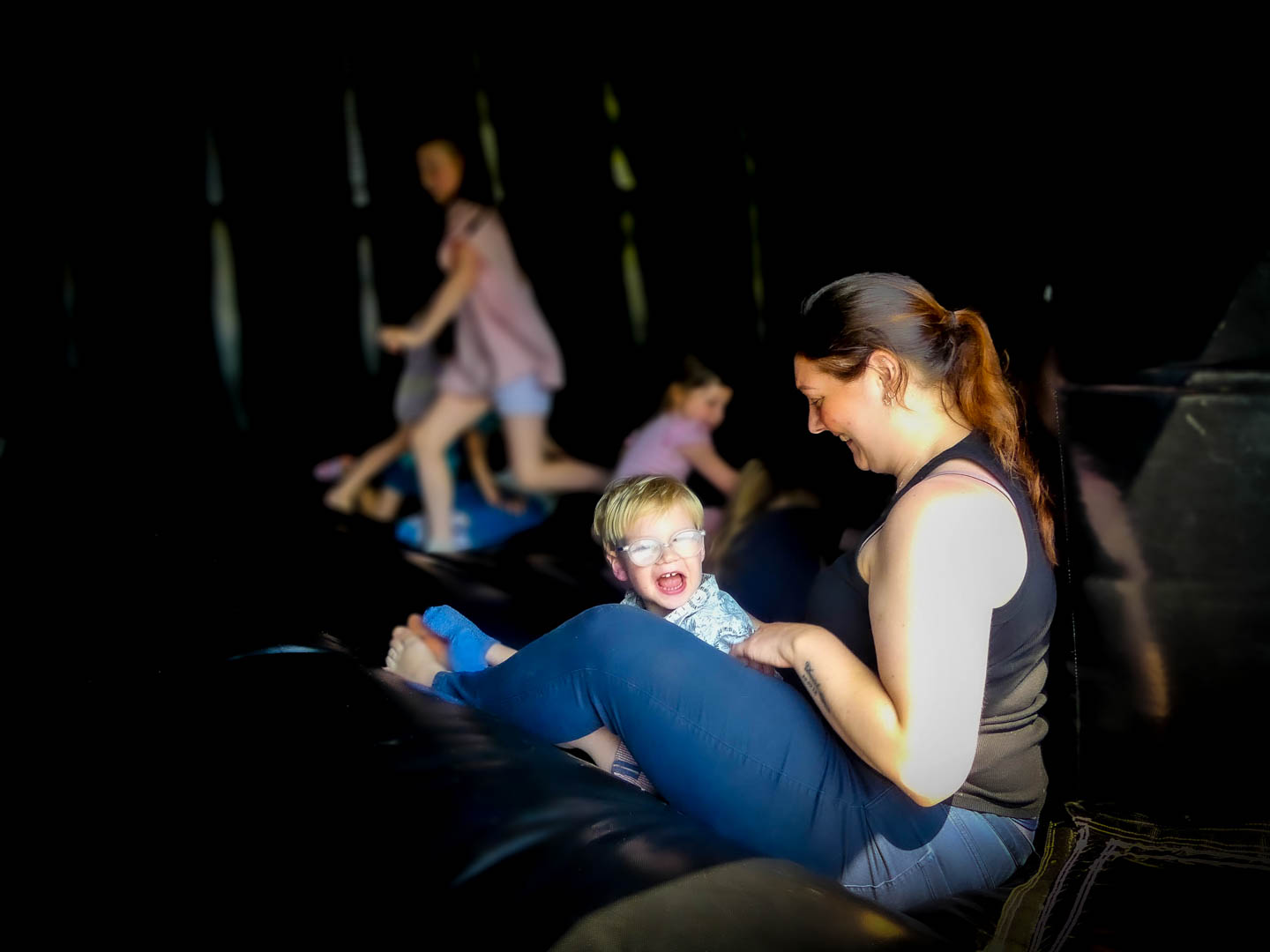 A woman with tied back hair sits on an inflatable structure, smiling at a young child sitting beside her who is wearing glasses and laughing. In the background, another child plays while an adult looks on. The scene is dimly lit, highlighting the interaction.