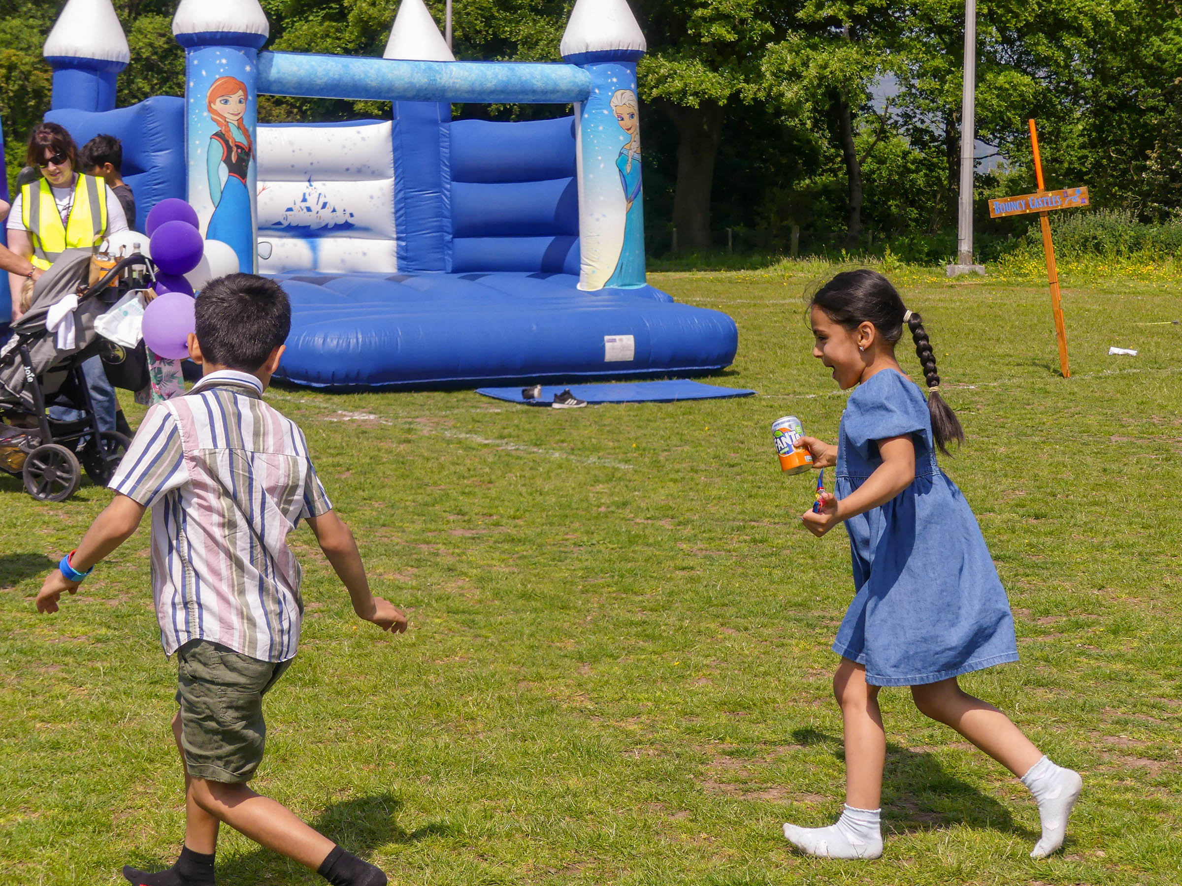 Two children play on a grassy field near a blue and white bouncy castle with animated princess characters on it. One child, wearing a plaid shirt and green shorts, runs towards the castle, while a girl in a blue dress holding a drink follows. Trees are in the background.