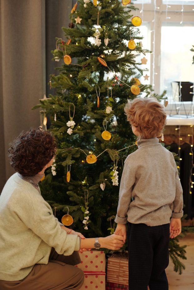 An adult and a child, both with curly hair, are seen decorating a Christmas tree with ornaments and dried orange slices. Caring at Christmas