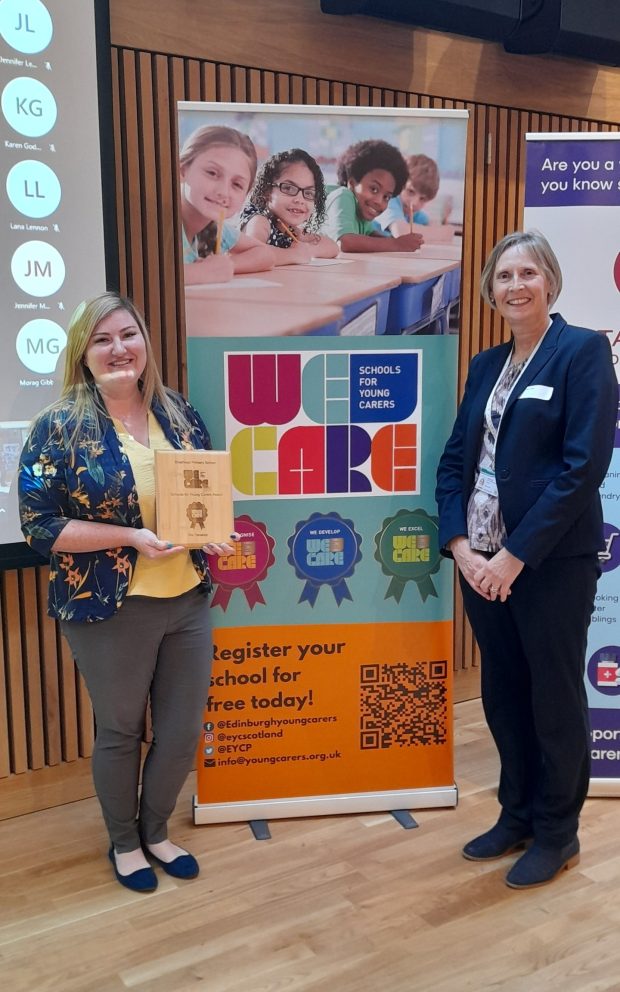 Two women, one holding a wooden award, stand in front of a We Care banner. Schools for Young Carers