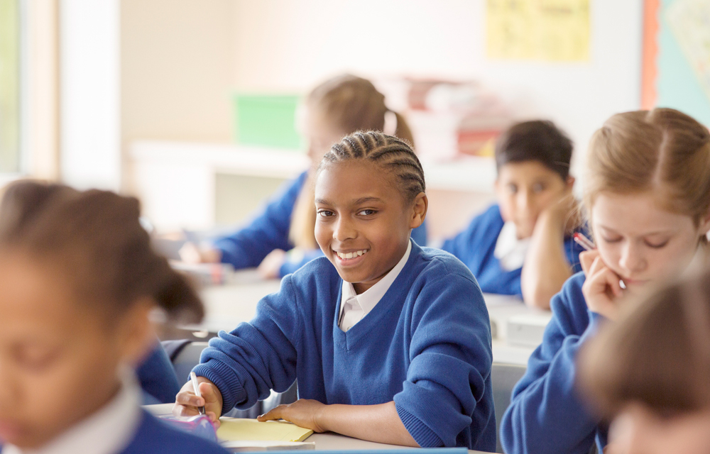 Students in blue uniforms sitting at desks in a classroom; one student is smiling at the camera.