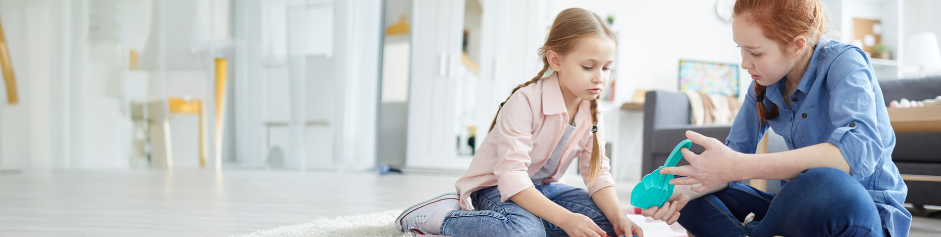 Two girls sitting on the floor, playing with toys in a bright living room.