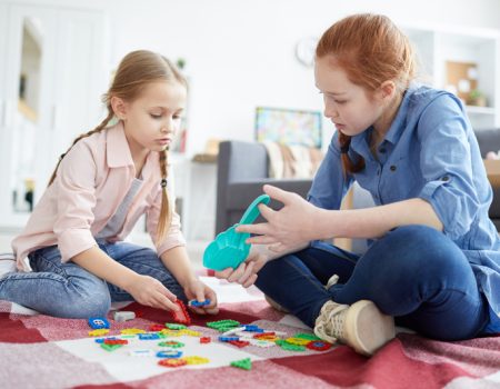 Two girls sitting on a red and white blanket, playing with colorful building blocks indoors.
