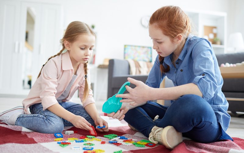 Two girls sitting on a red and white blanket, playing with colorful building blocks indoors.