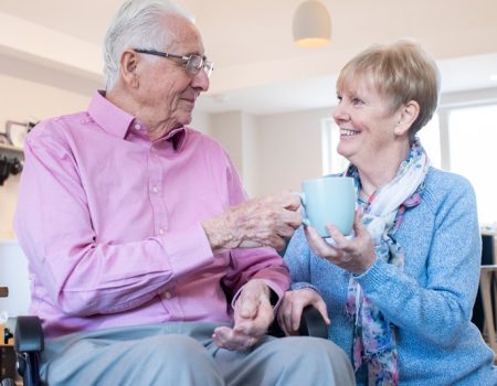 Older couple smiling, seated indoors. The man in a pink shirt holds a cup, while the woman, in blue, smiles at him.