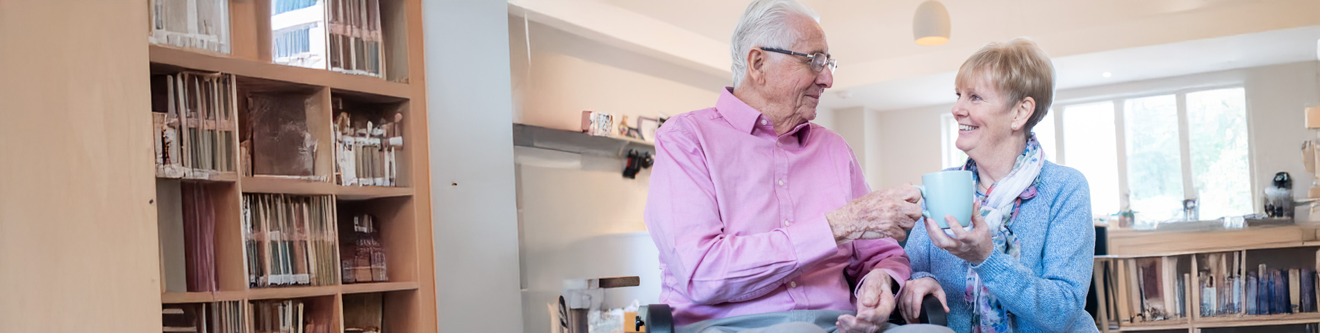 Elderly couple sharing tea and smiling in a cozy room with bookshelves.