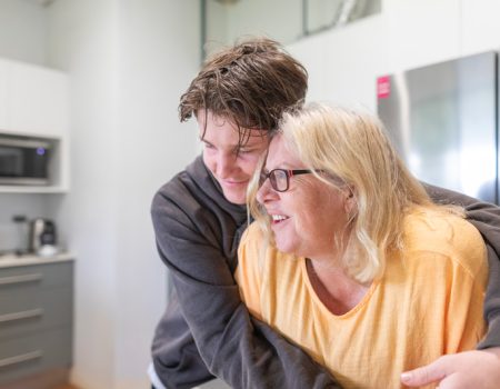 A young person hugs an older person in a kitchen, both smiling warmly.