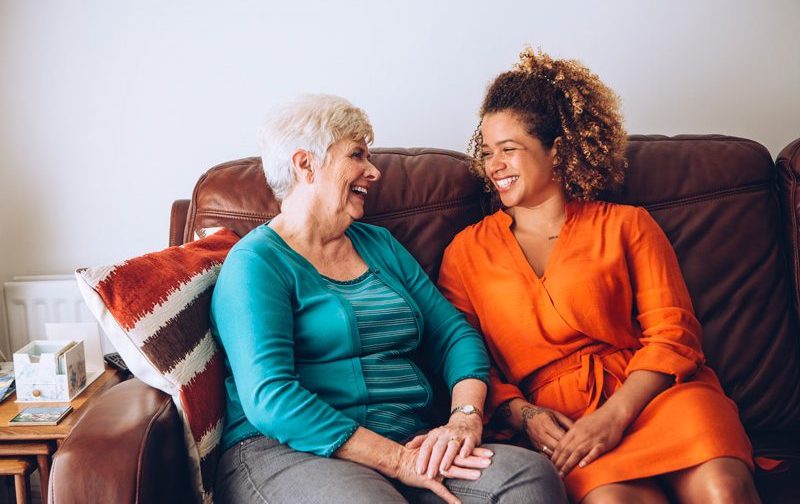 Two women sitting on a couch, smiling at each other. One is older with gray hair, the other has curly hair and wears orange.