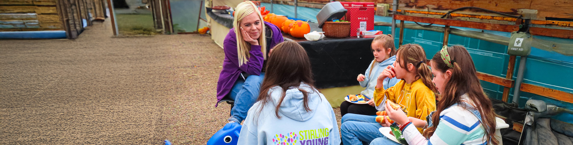 Group of children and a woman sitting in a rustic setting with pumpkins and snacks on a table.