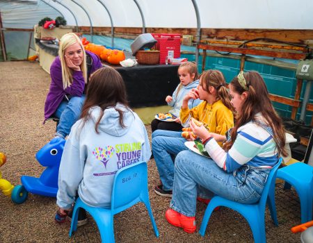 A group of children and an adult sit together in a greenhouse, having a conversation.