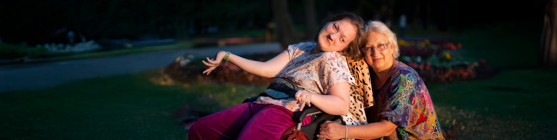 Woman in a wheelchair and another woman smiling in a park during twilight.