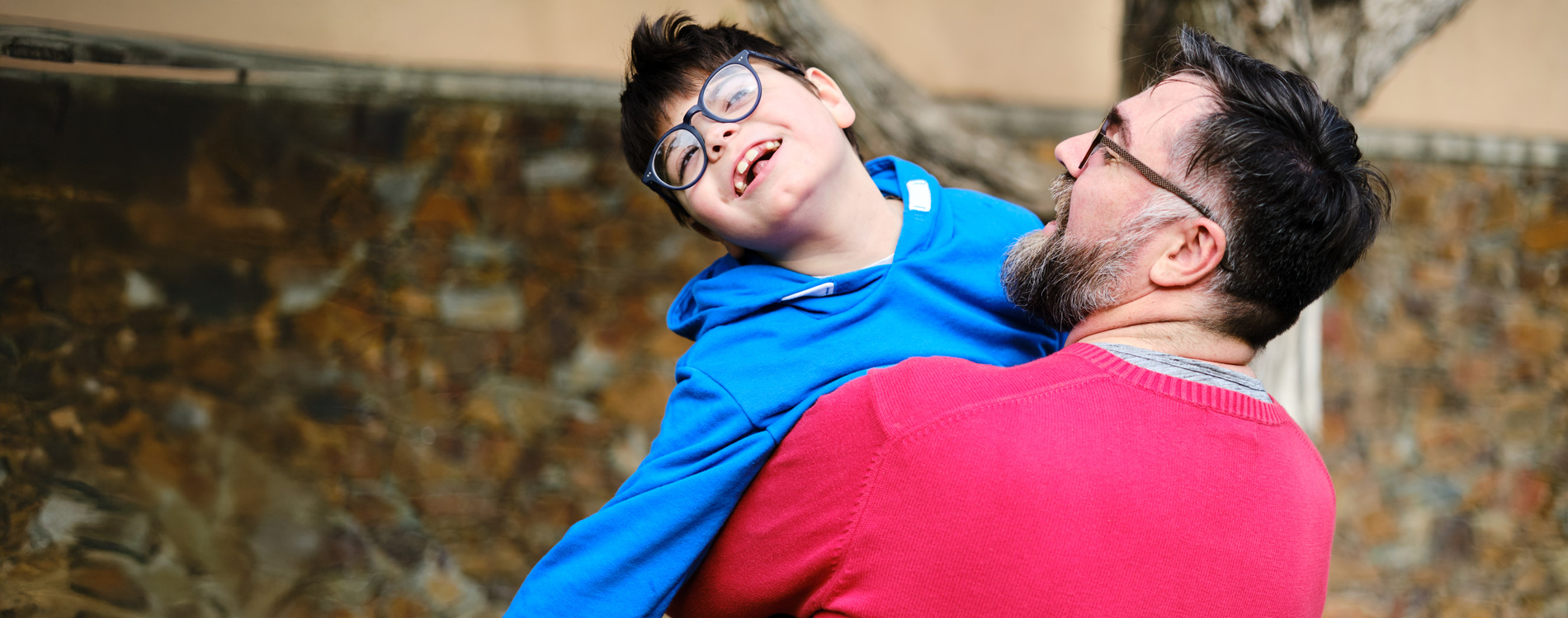 Man in red sweater lifting a smiling boy in blue hoodie, both wearing glasses, with a blurred outdoor background.