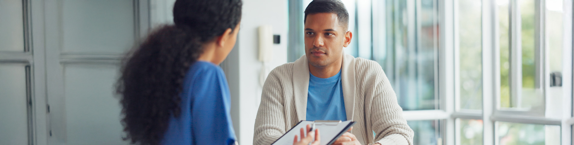 Two people having a serious conversation at a table in a bright room. One holds a clipboard.
