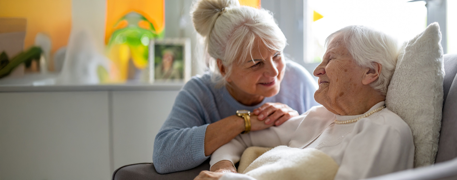 Elderly woman in bed smiles at a woman leaning over her, both appear content in a cozy room.