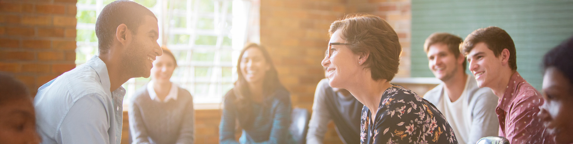 A diverse group of people smiling and talking in a bright, informal setting with sunlight streaming in from a window.