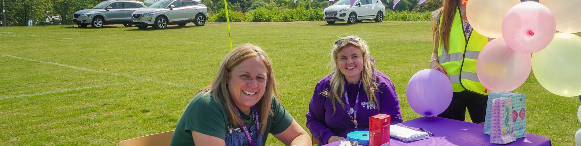 Three people at a purple table with balloons, outdoors on a sunny day, with cars and trees in the background.