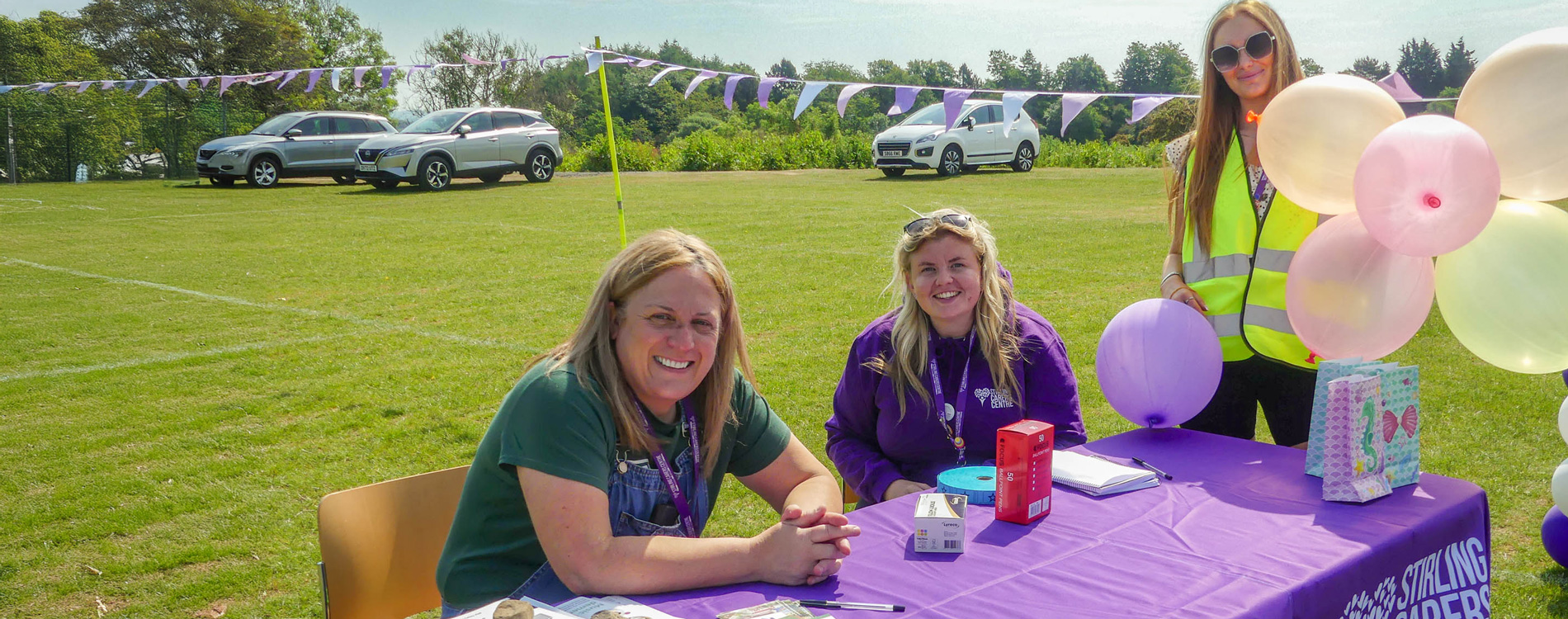 Three people at a purple table with balloons, outdoors on a sunny day, with cars and trees in the background.